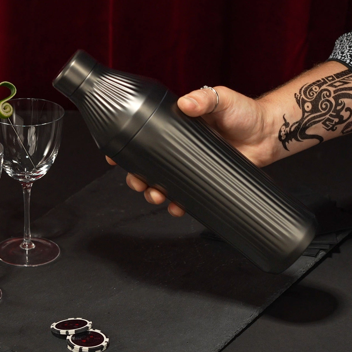 Hand holding Gunmetal Black Elevated Craft Diamond Edition Hybrid Cocktail Shaker with tattoos on arm in front of nick and nora glasses with lime peel garnish