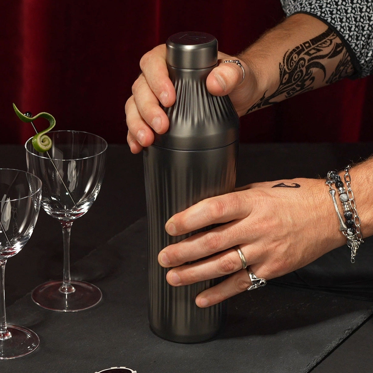 Man&#39;s hand opening Gunmetal Black Elevated Craft Diamond Edition Hybrid Cocktail Shaker measuring top next to nick and nora glasses with lime peel garnish