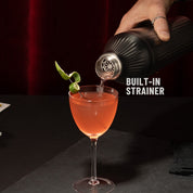 Hand pouring liquid through strainer of Gunmetal Black Elevated Craft Diamond Edition Hybrid Cocktail Shaker into Nick and Nora glass with lime peel garnish