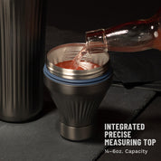 Bottle pouring red liquid into Gunmetal Black Elevated Craft Diamond Edition Hybrid Cocktail Shaker measuring top next to tumbler