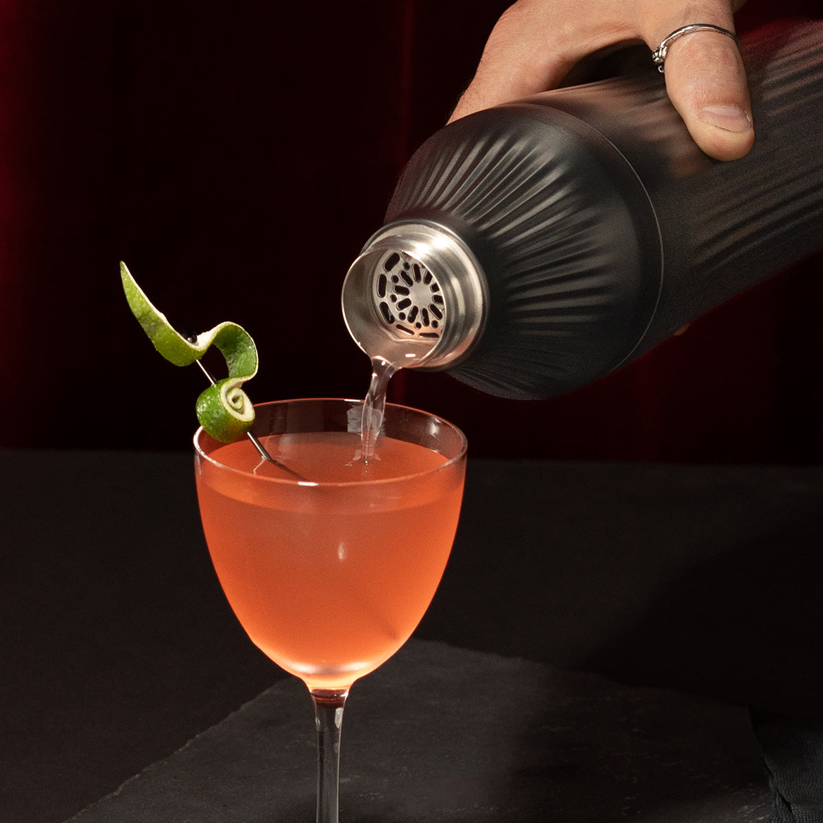 Hand pouring liquid through strainer of Gunmetal Black Elevated Craft Diamond Edition Hybrid Cocktail Shaker into nick and nora glass with lime peel garnish