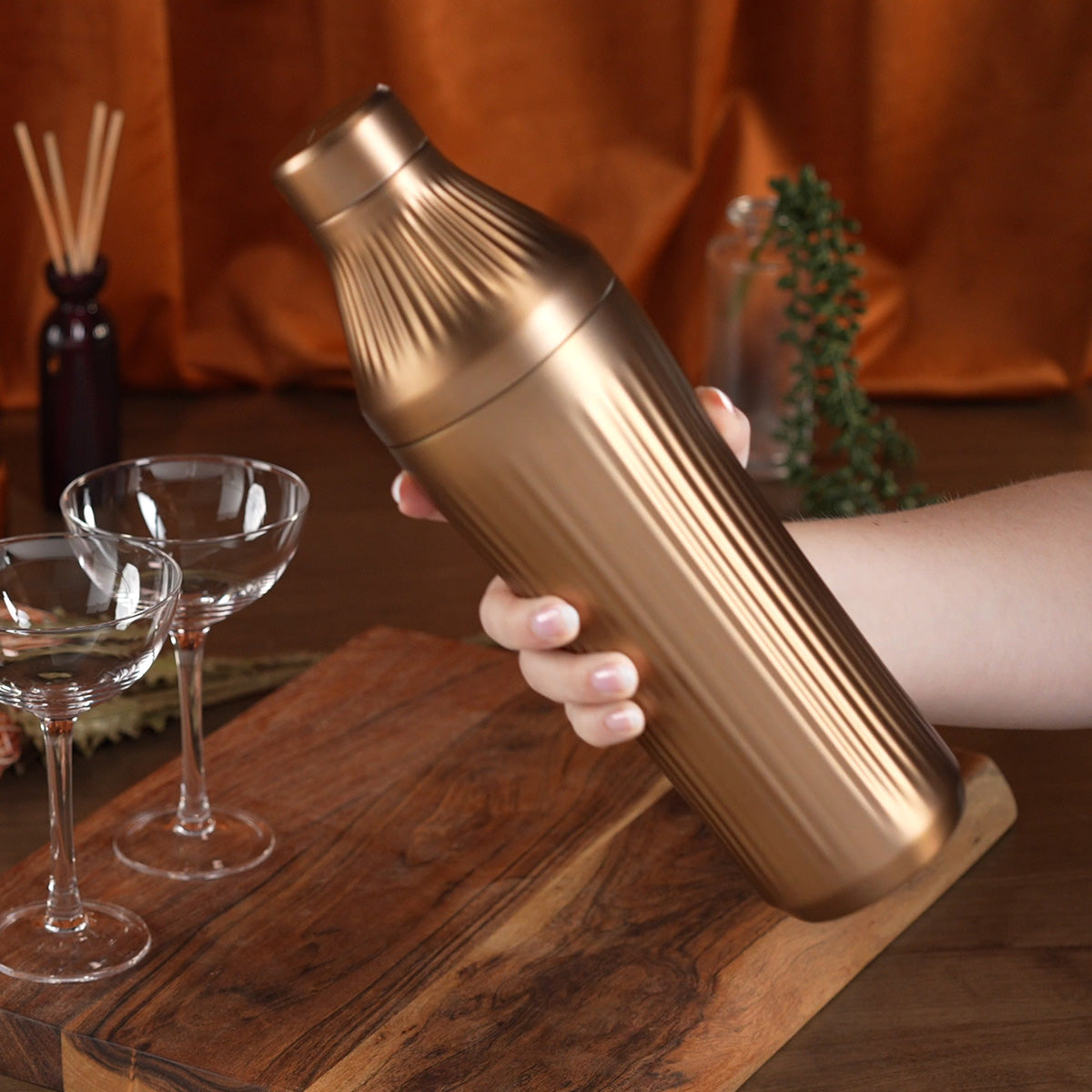 Hand holding Brushed Copper Elevated Craft Diamond Edition Hybrid Cocktail Shaker in front of empty couple glasses