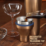 Bottle pouring brown liquid into Brushed Coper Elevated Craft Diamond Edition Hybrid Cocktail Shaker measuring top next to tumbler and empty coupe glass