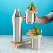 Hand holding the Elevated Craft Brushed Stainless Hybrid Cocktail Glass filled with light yellow cocktail topped with mint and an orange umbrella next to the Hybrid Cocktail Shaker