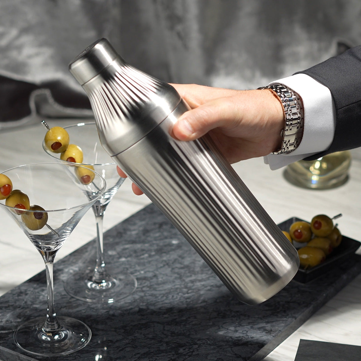 Man in suit&#39;s hand holding Stainless Steel Elevated Craft Diamond Edition Hybrid Cocktail Shaker in front of martini glasses with olives