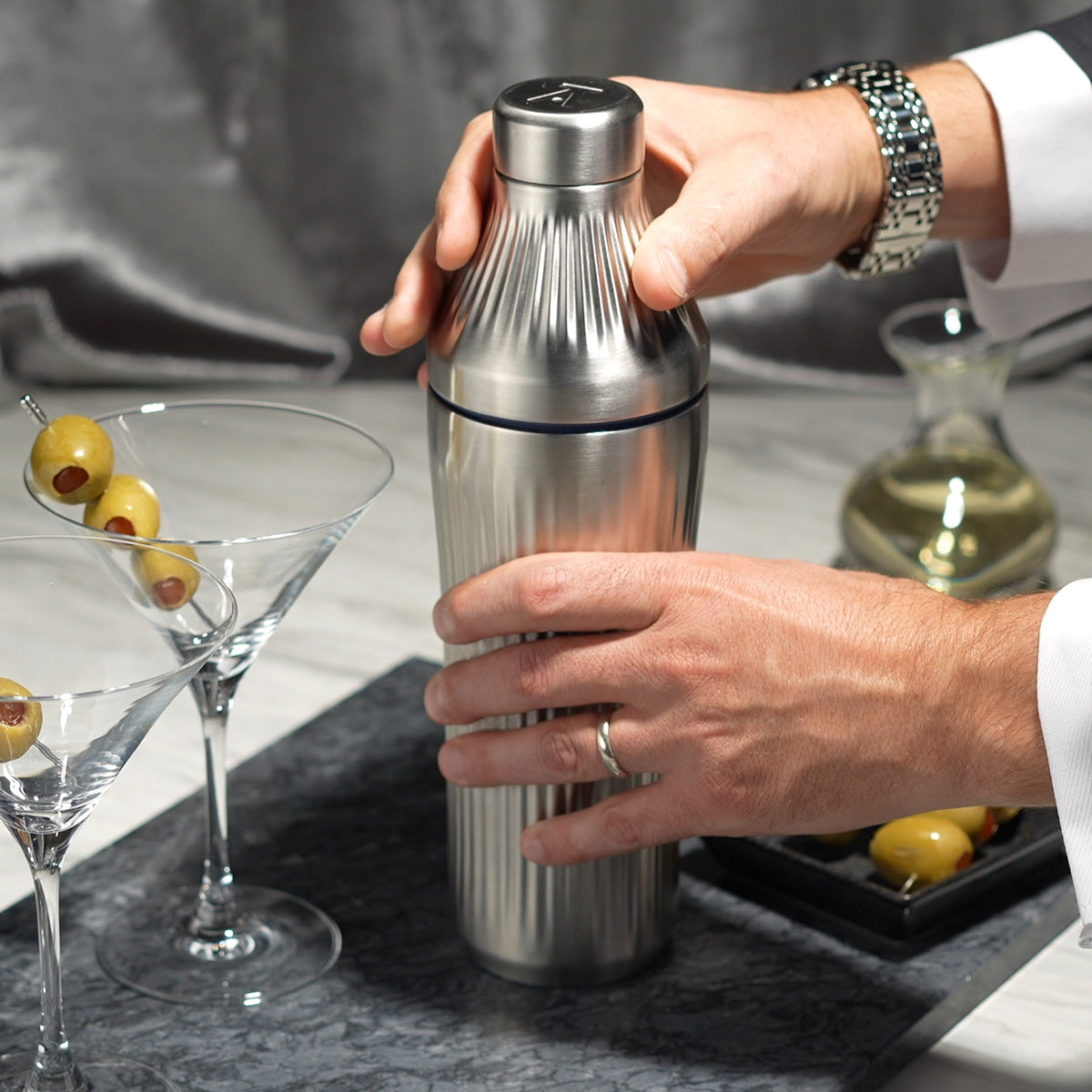 Man in suit&#39;s hand opening Stainless Steel Elevated Craft Diamond Edition Hybrid Cocktail Shaker measuring top next to martini glass with olives