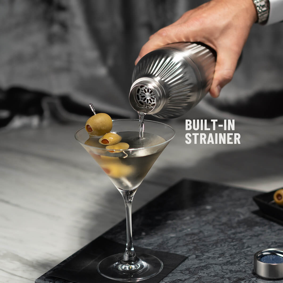 Hand pouring liquid through strainer of Stainless Steel Elevated Craft Diamond Edition Hybrid Cocktail Shaker into martini glass with olives
