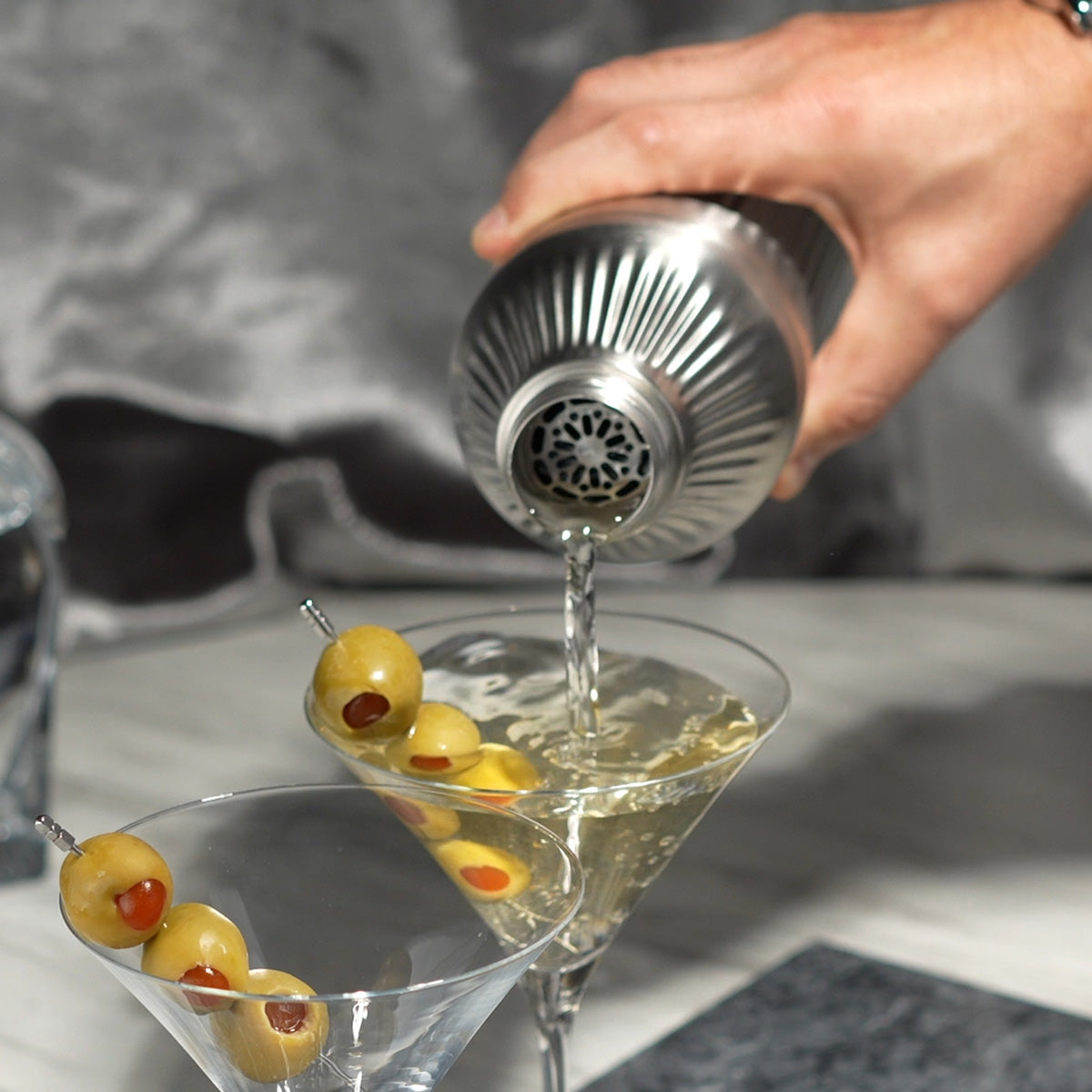 Hand pouring liquid through strainer of Stainless Steel Elevated Craft Diamond Edition Hybrid Cocktail Shaker into martini glass with olives