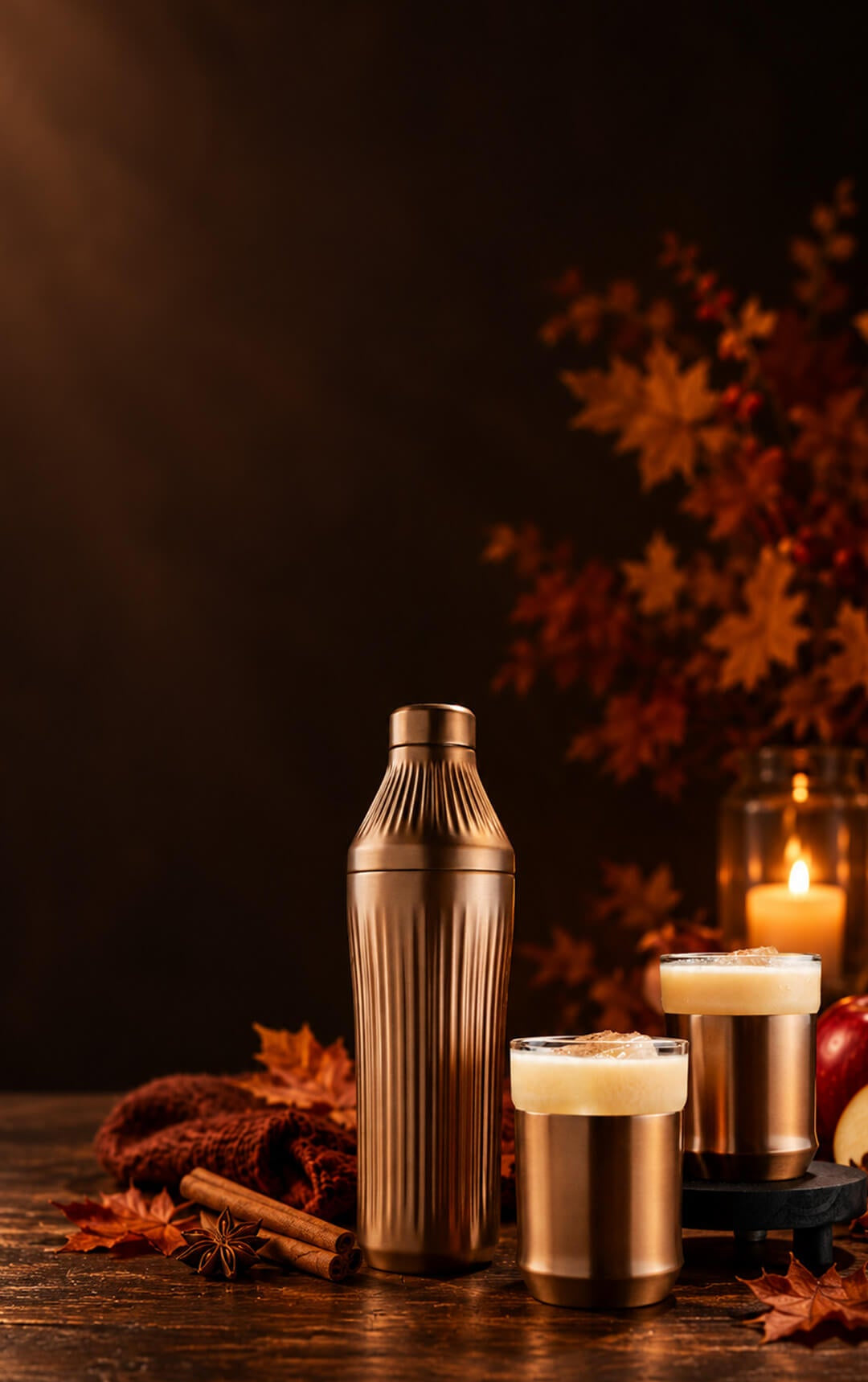 A metallic bronze cocktail shaker and two matching cups arranged on a dark wooden table with autumn leaves, cinnamon sticks, and a lit candle in the background.