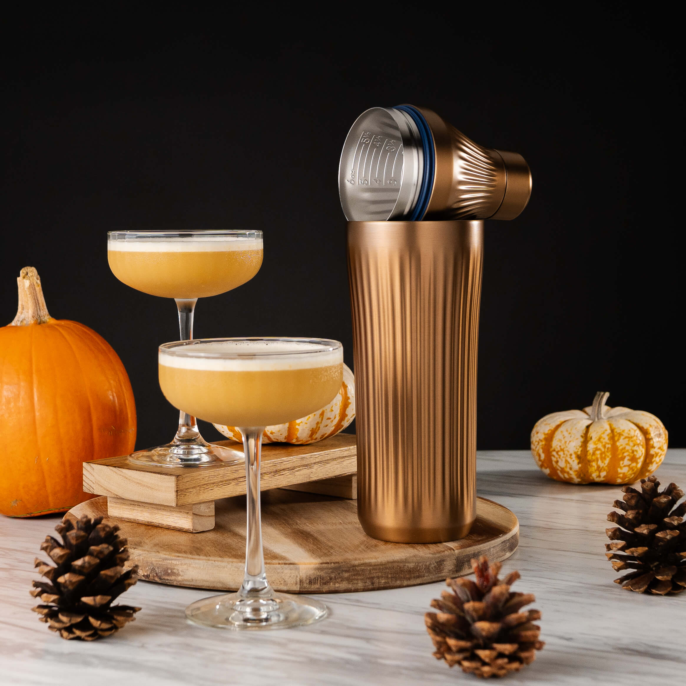 Copper Cocktail Shaker next to two glasses of a fall drink with pumpkin and pinecones on a table next to it