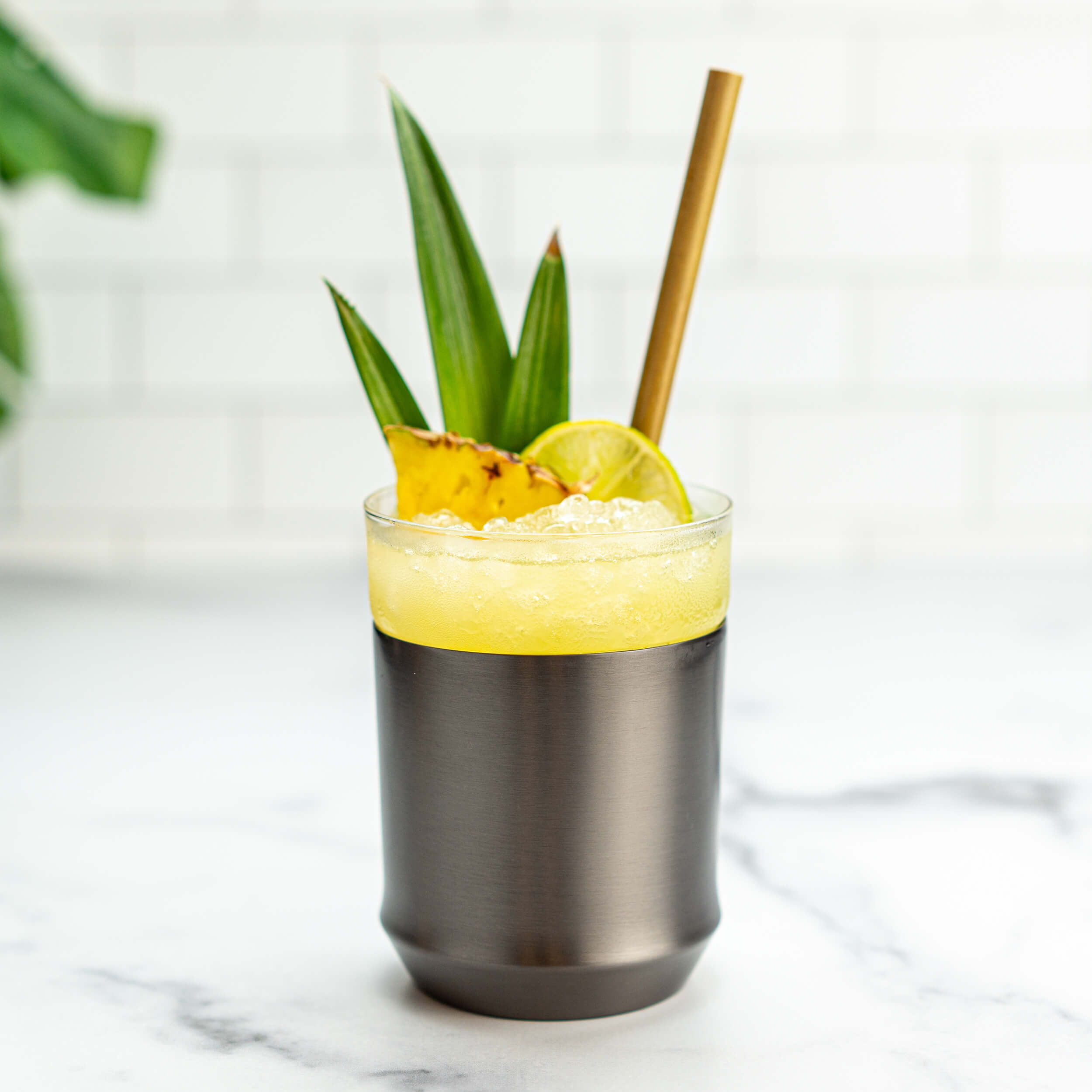 The Elevated Craft Hybrid Cocktail Glass in gunmetal black finish filled with a bright yellow tiki cocktail with pebble ice garnished with pineapple fronds a pineapple chunk and a lime wheel