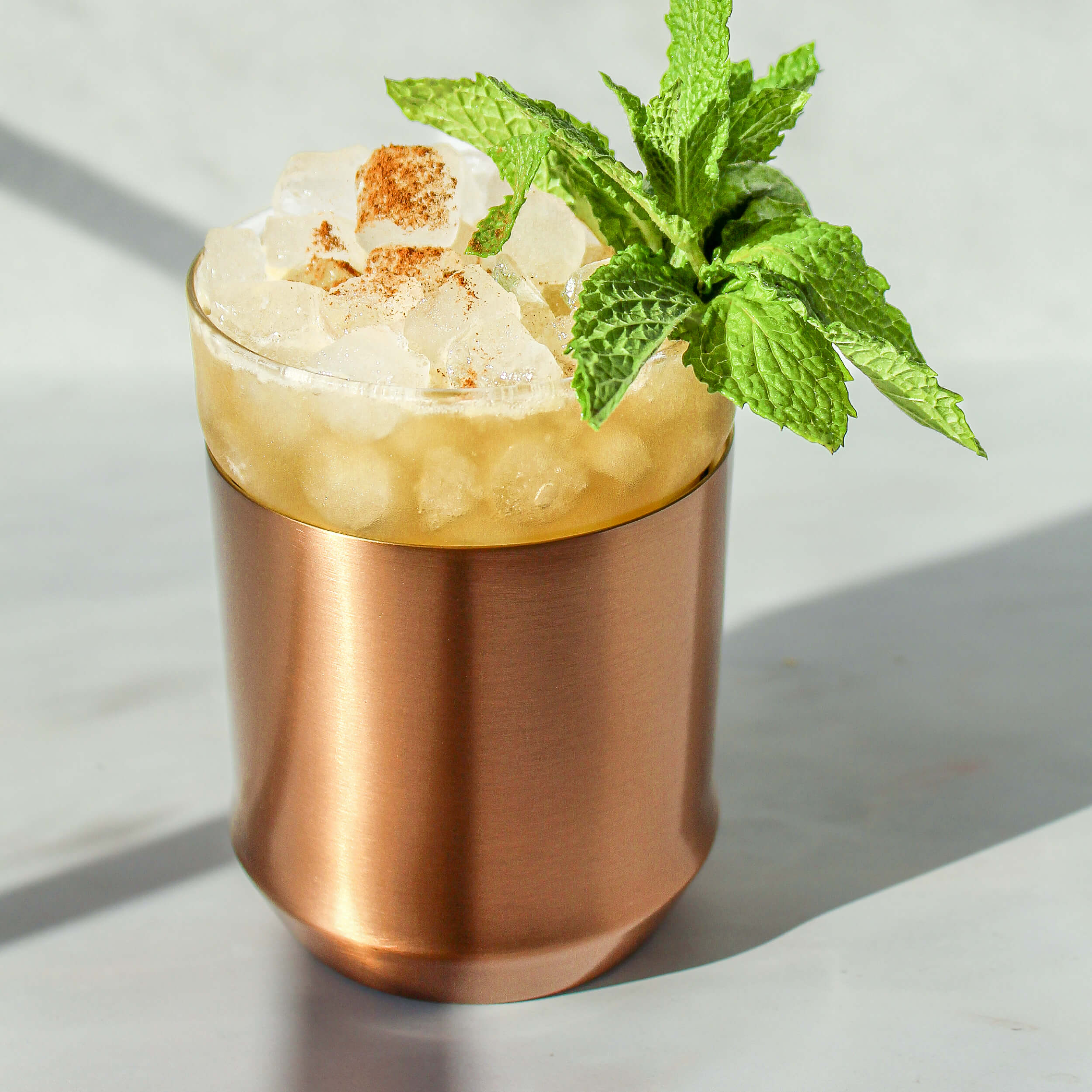 The Elevated Craft Hybrid Cocktail Glass in brushed copper finish filled with a bright yellow cocktail in pebble ice garnished with a full mint sprig and freshly hand-grated nutmeg