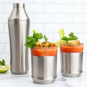 Hybrid Cocktail Shaker & two Hybrid Cocktail Glasses in brushed stainless finish filled with bloody mary cocktails garnished with lime wedges green olives celery sticks and parsley