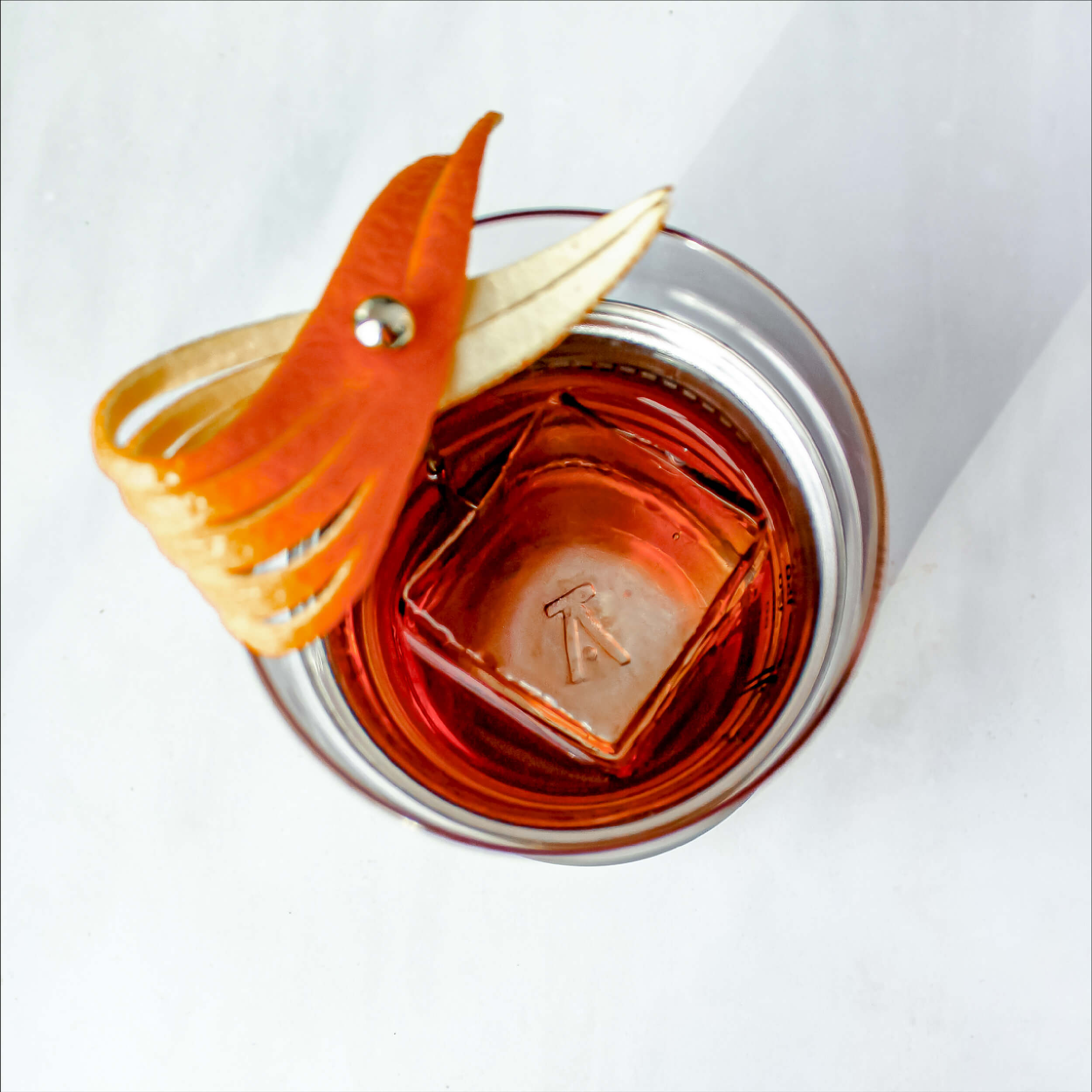 Stainless steel Hybrid Cocktail Glass 
 viewed from above filled with a dark orange cocktail and single large ice cube garnished with an orange peel