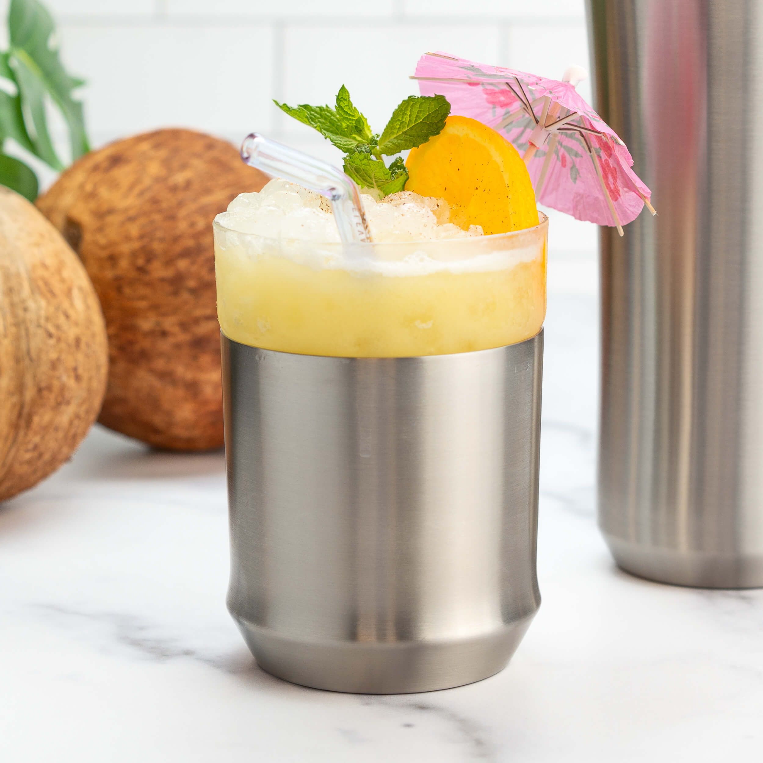 Elevated Craft Hybrid Cocktail Glass in Brushed Stainless finish filled with a bright yellow tiki cocktail in pebble ice garnished with mint orange slice and miniature umbrella