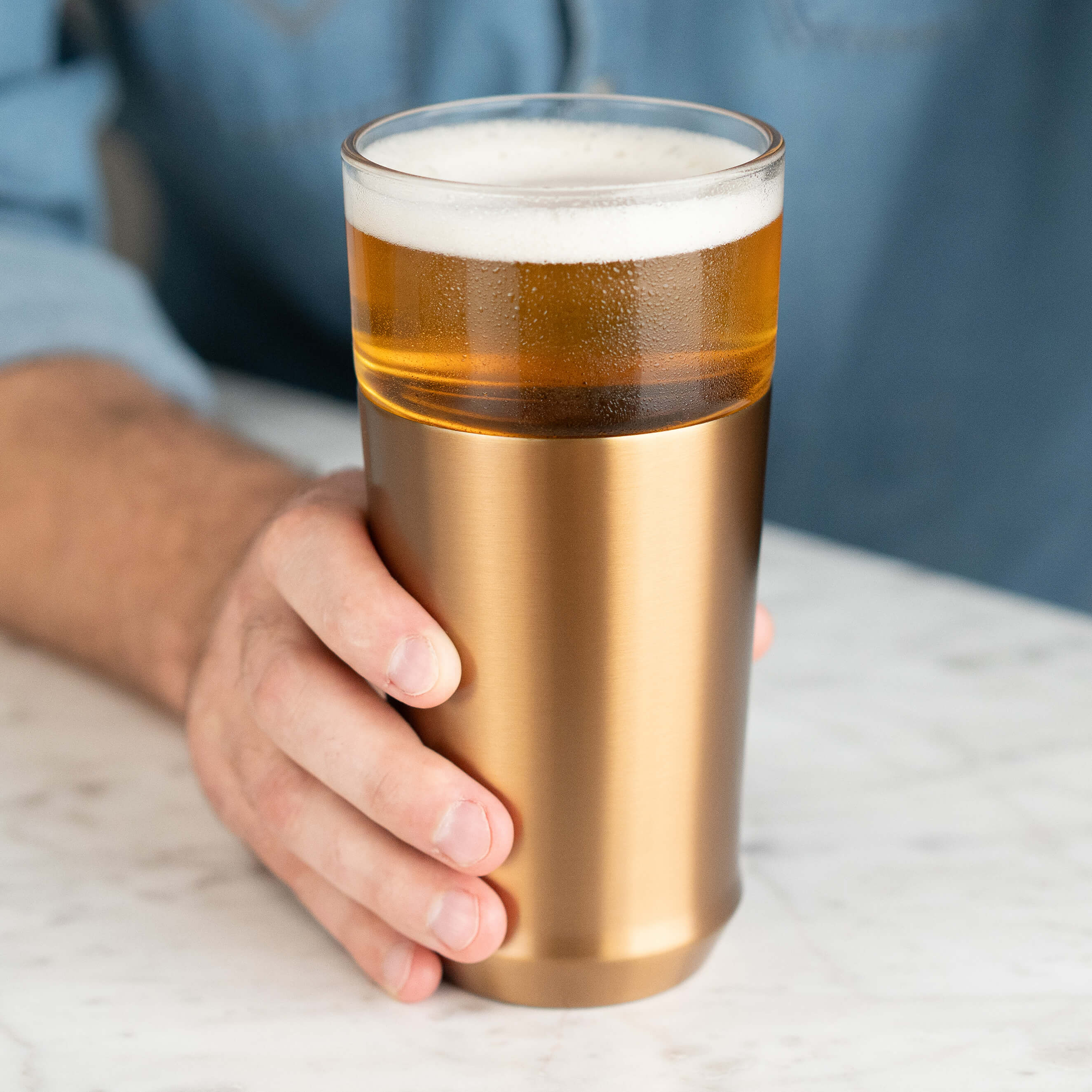 A man holding the Elevated Craft Hybrid Pint Glass in the Brushed copper finish filled with chilled full bodied beer on a marble surface