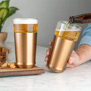 Person pouring cold beer from a bottle into the Elevated Craft Hybrid Pint Glass in brushed copper finish next to another brushed copper finish hybrid pint glass full of cold beer