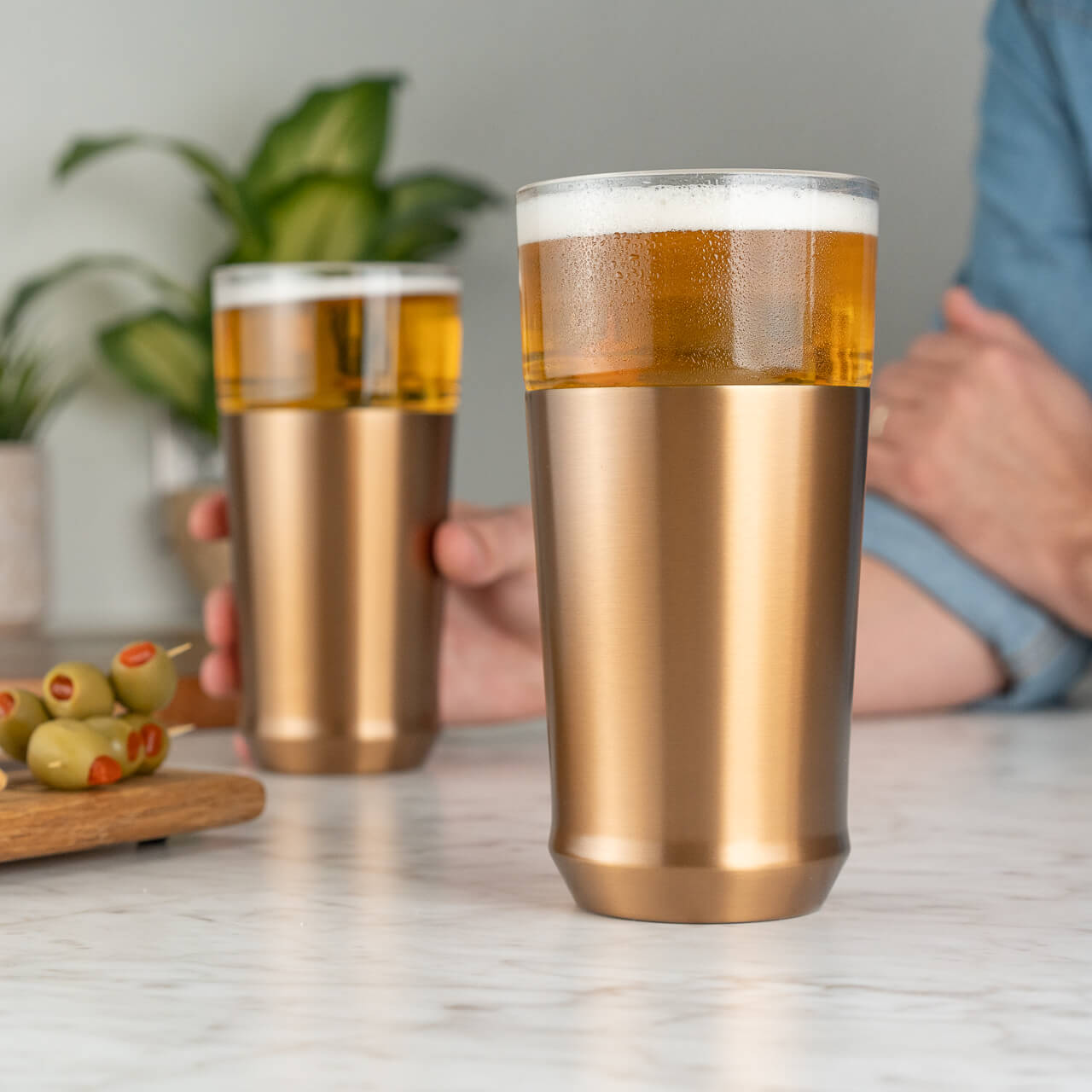 Elevated Craft Hybrid Pint Glass in brushed copper finish full of cold beer in foreground with a hand holding another brushed copper finish hybrid pint glass full of cold beer in the background