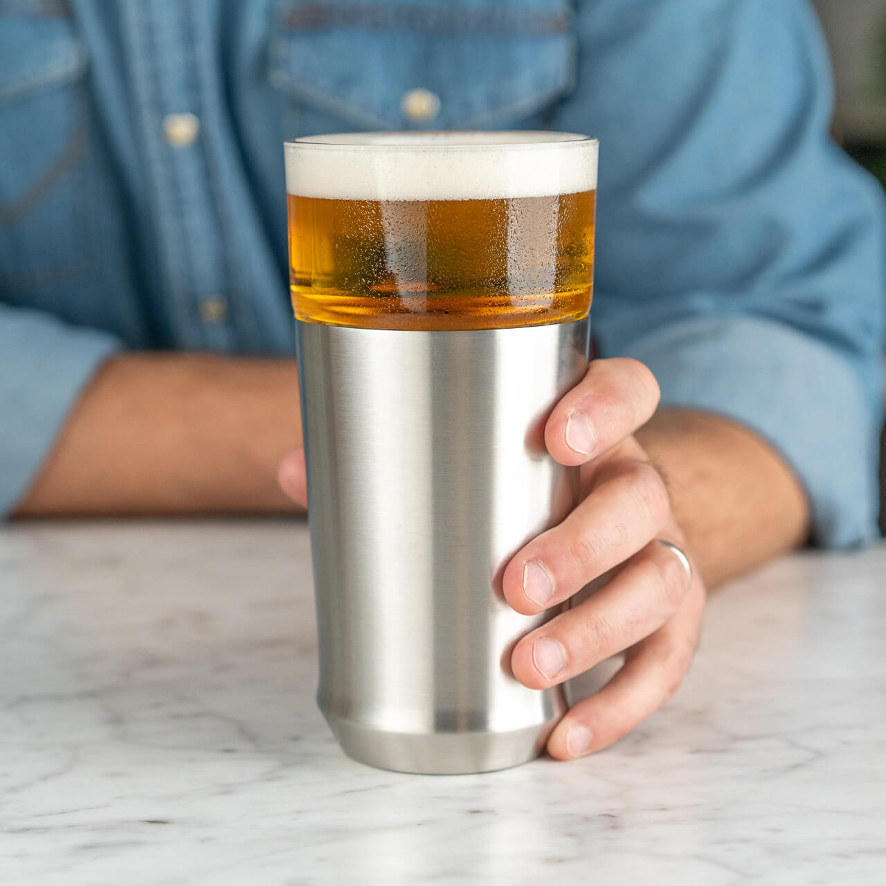 A man holding the Elevated Craft Hybrid Pint Glass in the Brushed Stainless finish filled with chilled full bodied beer on a marble surface