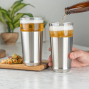 Man pouring cold beer from a bottle into the Elevated Craft Hybrid Pint Glass in brushed stainless finish next to another stainless hybrid pint glass full of cold beer