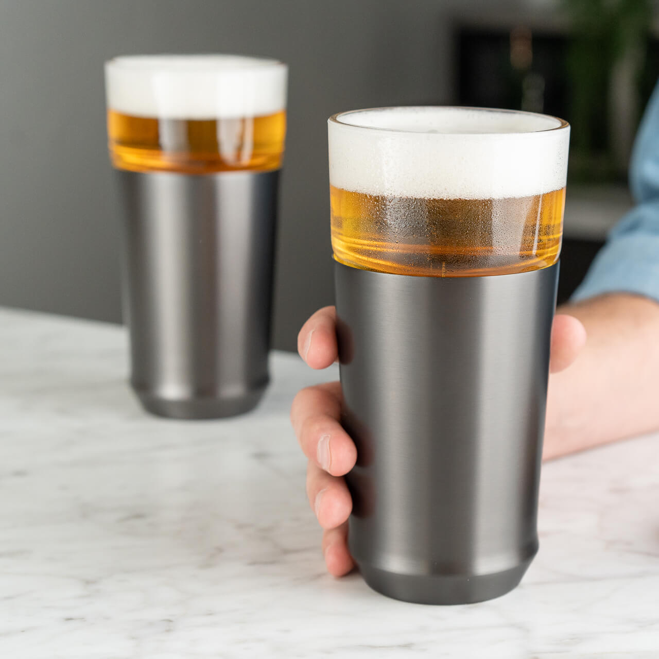 The Elevated Craft hybrid pint glass in gunmetal black finish full of cold beer being held by a hand over a marble countertop in the foreground with another hybrid pint glass full of cold beer in the background