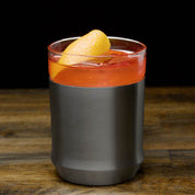 The Elevated Craft hybrid cocktail glass in gunmetal black finish filled with an amber cocktail large ice cube and orange peel garnish
