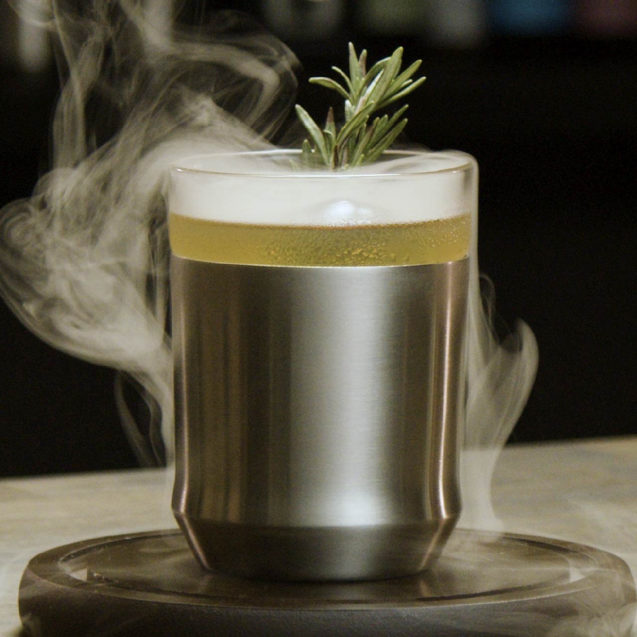 The Elevated Craft Hybrid Cocktail Glass in brushed stainless finish filled with a smoking yellow cocktail garnished with a rosemary sprig