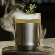 The Elevated Craft Hybrid Cocktail Glass in brushed stainless finish filled with a smoking yellow cocktail garnished with a rosemary sprig