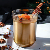 The Elevated Craft Hybrid Cocktail Glass in brushed copper finish filled with a dark orange cockail garnished with a cinnamon stick, muddled rasberries orange peel and star anise balanced on the glass insert rim